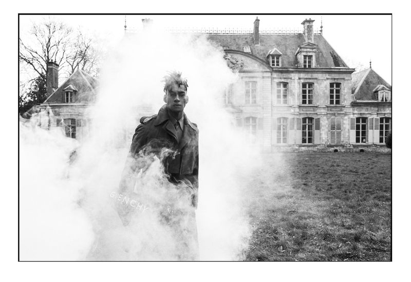 Homme noir en trench Givenchy émergeant d’un nuage de fumée devant un château